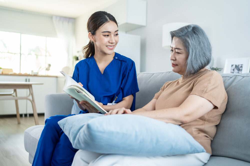 Asian woman nurse taking care of senior elderly female. Young Caregiver Therapist doctor sit on sofa, read a book to happy older handicapped patient.
