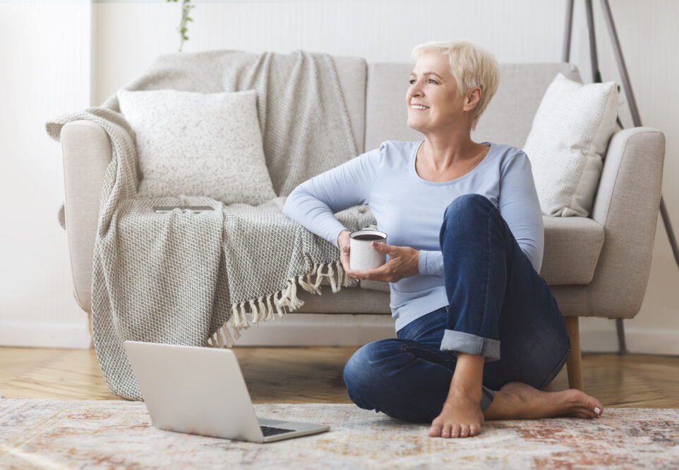 Cheerful elderly business lady sitting on floor at home, drinking coffee, using laptop, free space. Independent Living.