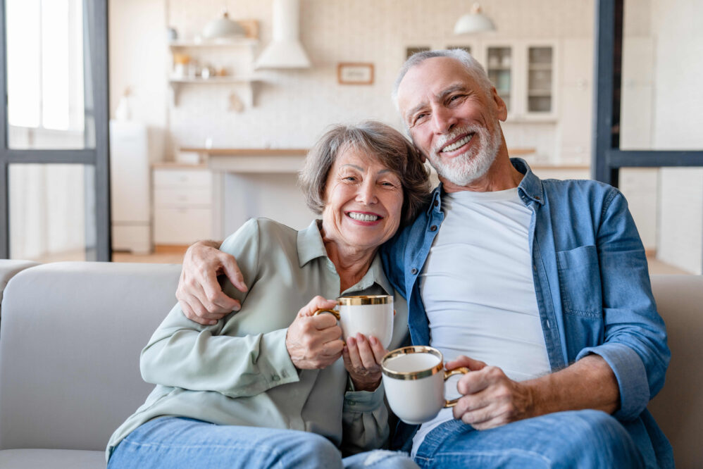 A cheerful old senior happy couple drinking hot beverage tea while looking at camera sitting on the sofa