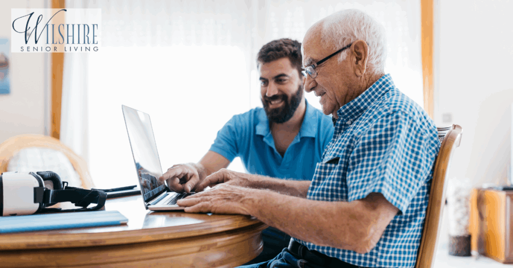An elderly man smiles while using a laptop at a table, assisted by a younger man, possibly a family member, in a bright room at a memory care community.