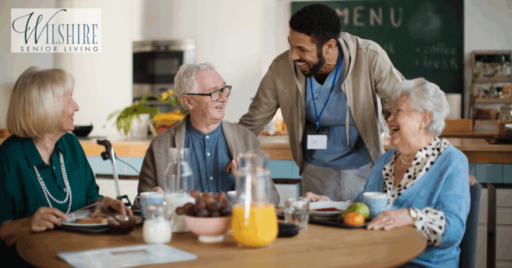 A group of older adults share a joyful breakfast at a memory care community dining area, smiling and engaging with a friendly care partner wearing a name badge. The atmosphere is lively and welcoming.